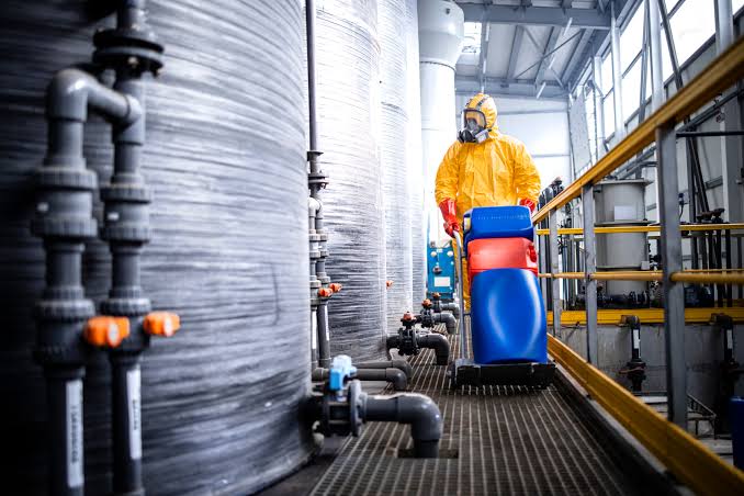 Industrial worker wearing a protective hazmat suit near large chemical storage tanks at a manufacturing plant