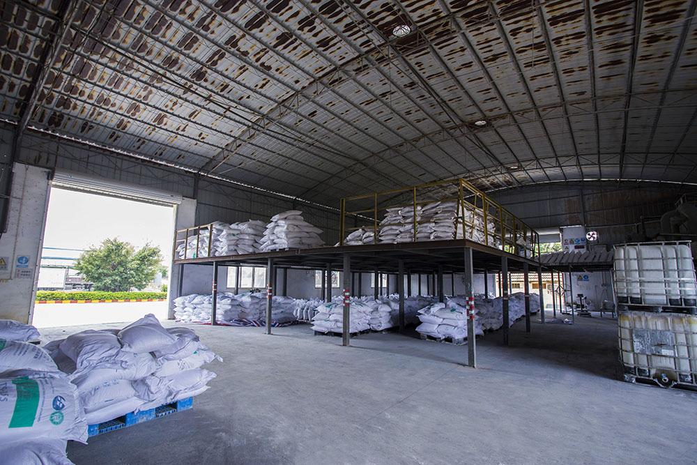 Large industrial chemical storage warehouse with stacked white pallets of bagged product and intermediate bulk container (IBC) totes under a high corrugated roof