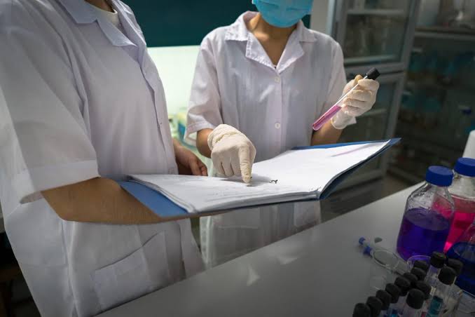 Two laboratory technicians in white coats reviewing test results on a binder while holding a sample test tube
