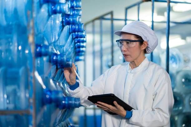 A professional Asian female quality control inspector in a white coat and safety gear conducting an audit of a modern chemical production facility