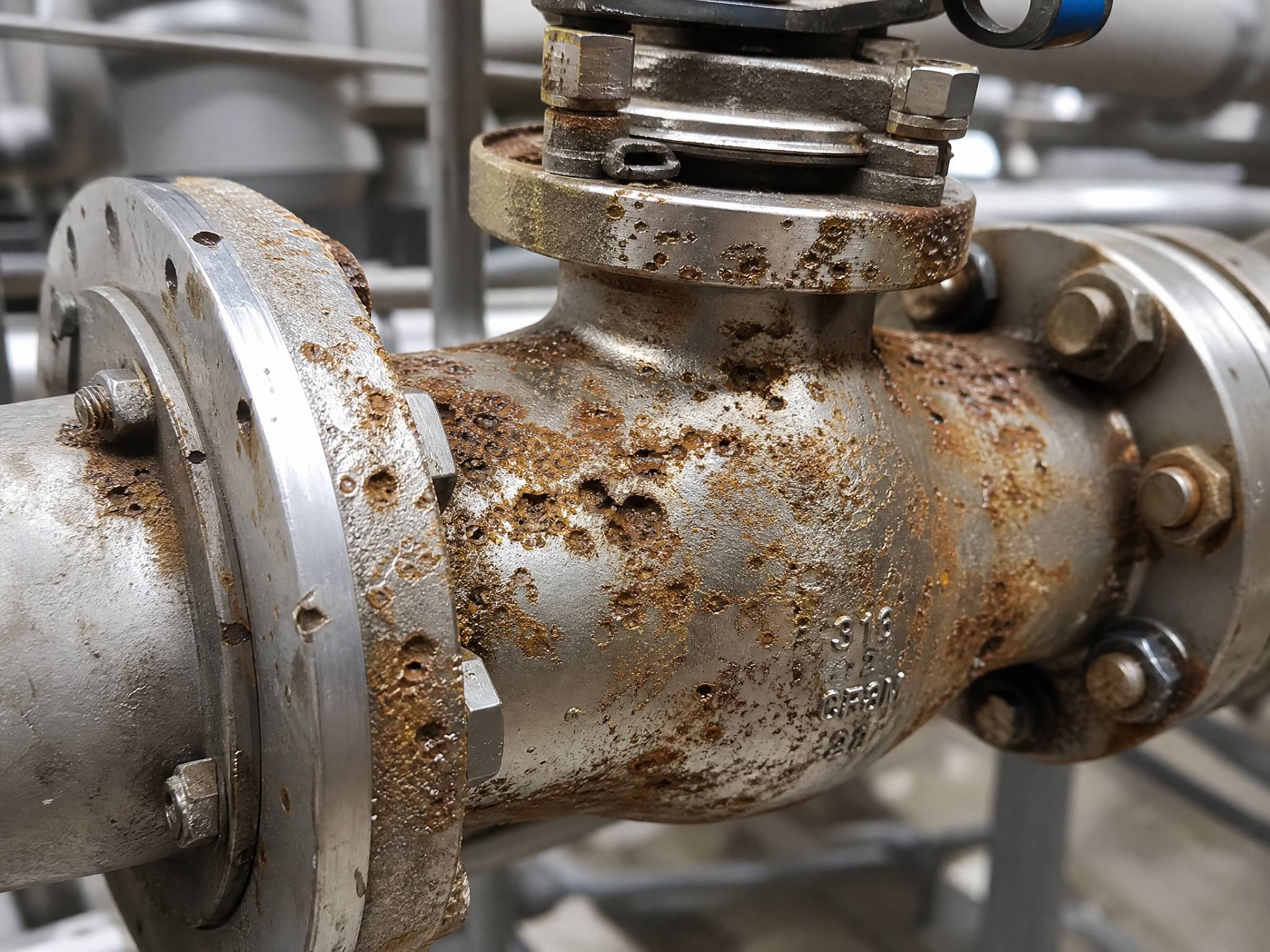 A close-up of a stainless steel valve or pipe showing significant pitting and rust-colored corrosion. The surface is uneven and damaged. Suggests the negative impact of using low-quality chemicals. Industrial, detailed, photorealistic.