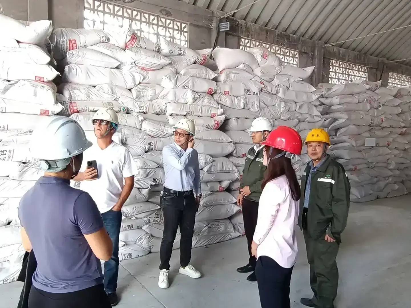 Industrial workers and business professionals in safety helmets discussing a purchase order in a chemical warehouse filled with stacked bulk bags