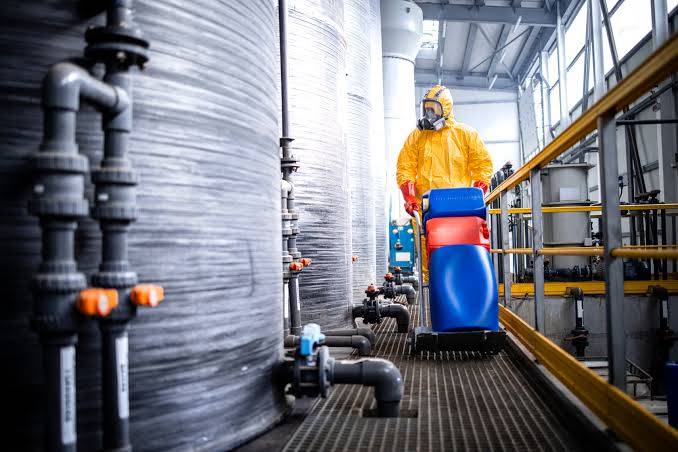 Industrial worker wearing a protective hazmat suit near large chemical storage tanks at a manufacturing plant